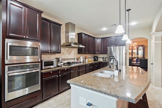 kitchen featuring wall chimney exhaust hood, stainless steel appliances, sink, decorative light fixtures, and an island with sink