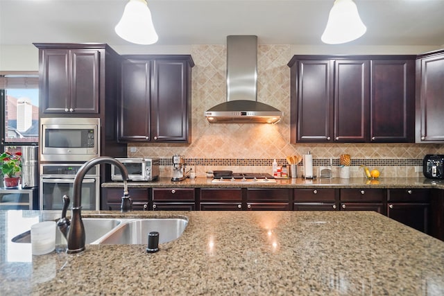 kitchen featuring light stone counters, wall chimney range hood, stainless steel appliances, and hanging light fixtures