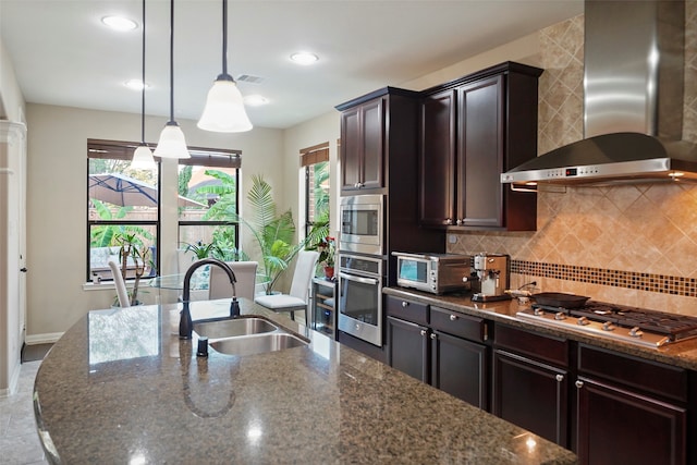 kitchen featuring sink, stainless steel appliances, wall chimney range hood, dark stone countertops, and decorative light fixtures