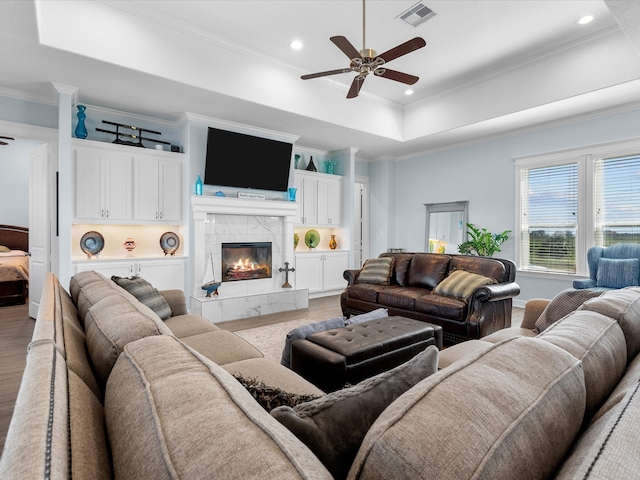 living room with ceiling fan, light hardwood / wood-style floors, a tiled fireplace, a tray ceiling, and ornamental molding