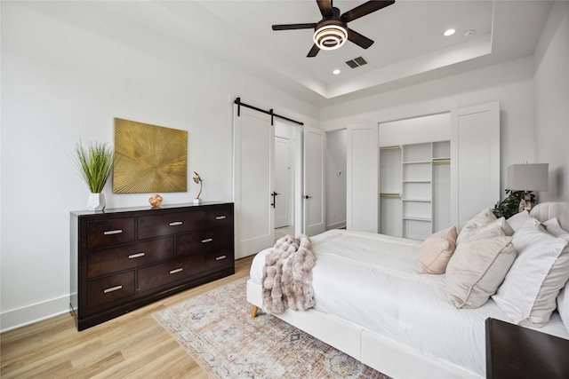 bedroom with a barn door, light hardwood / wood-style floors, a raised ceiling, and ceiling fan