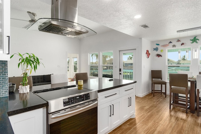 kitchen featuring light wood-type flooring, stainless steel range oven, island range hood, white cabinetry, and a textured ceiling