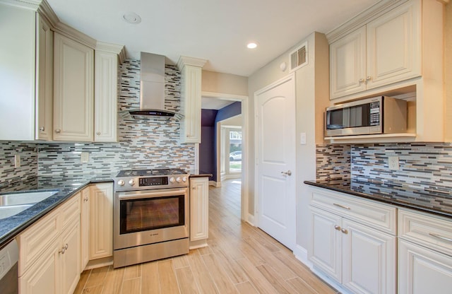 kitchen with stainless steel appliances, wall chimney range hood, backsplash, dark stone countertops, and cream cabinetry