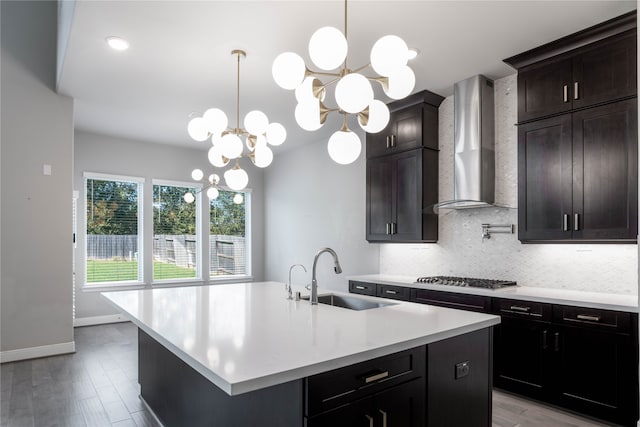 kitchen featuring wall chimney range hood, sink, light wood-type flooring, stainless steel gas cooktop, and a kitchen island with sink