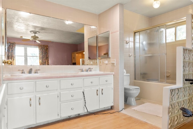 full bathroom with combined bath / shower with glass door, tile walls, vanity, ceiling fan, and toilet