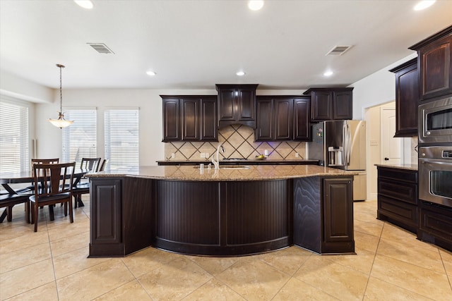 kitchen with a kitchen island with sink, dark brown cabinets, appliances with stainless steel finishes, light stone counters, and tasteful backsplash