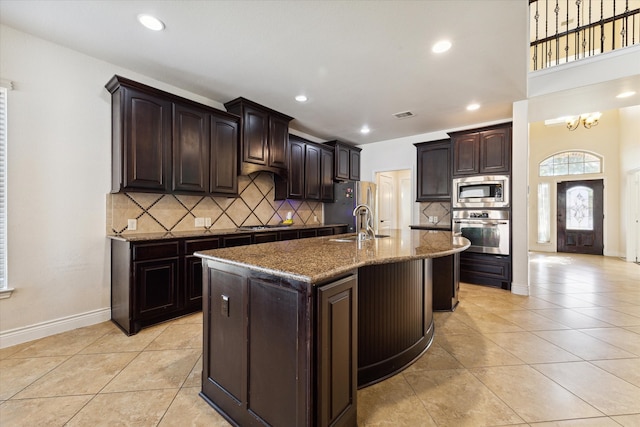 kitchen with backsplash, stainless steel appliances, dark brown cabinetry, light tile patterned floors, and a kitchen island with sink