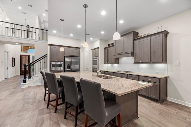 kitchen with appliances with stainless steel finishes, light wood-type flooring, a kitchen island with sink, sink, and decorative light fixtures