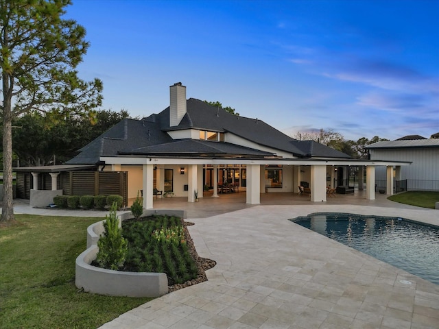 back of property at dusk featuring a patio area, a chimney, an outdoor pool, and a lawn