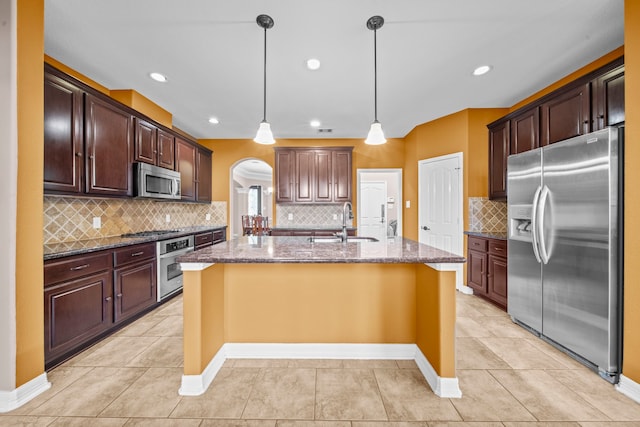 kitchen with an island with sink, stainless steel appliances, dark stone countertops, sink, and decorative light fixtures