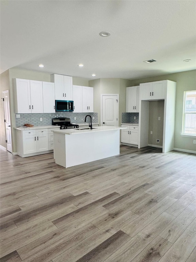 kitchen with an island with sink, white cabinetry, backsplash, stainless steel appliances, and light hardwood / wood-style flooring