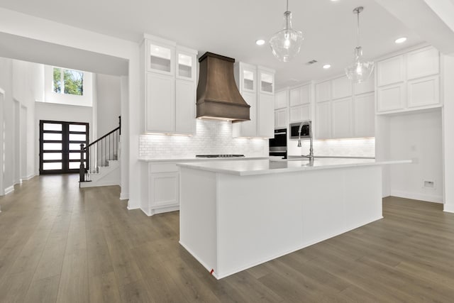 kitchen with white cabinetry, custom exhaust hood, an island with sink, appliances with stainless steel finishes, and hanging light fixtures