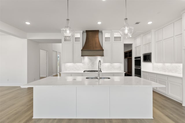 kitchen with custom exhaust hood, white cabinets, a kitchen island with sink, and light stone counters