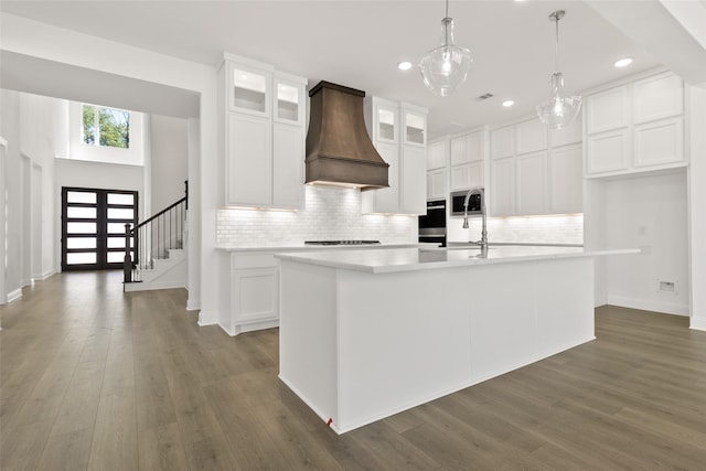 kitchen featuring stainless steel microwave, decorative light fixtures, premium range hood, a center island with sink, and white cabinetry