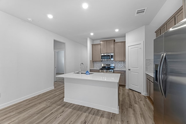 kitchen with light hardwood / wood-style floors, sink, tasteful backsplash, an island with sink, and appliances with stainless steel finishes