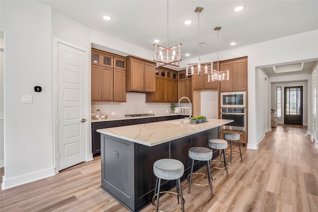 kitchen featuring light stone counters, decorative backsplash, a kitchen island with sink, light hardwood / wood-style flooring, and appliances with stainless steel finishes