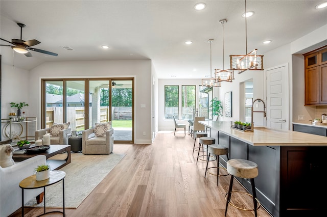 kitchen with light stone counters, an island with sink, hanging light fixtures, ceiling fan with notable chandelier, and light hardwood / wood-style floors