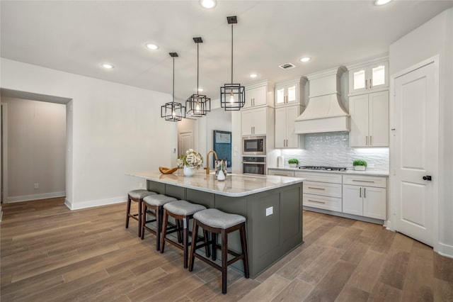 kitchen featuring an island with sink, premium range hood, stainless steel appliances, and white cabinetry