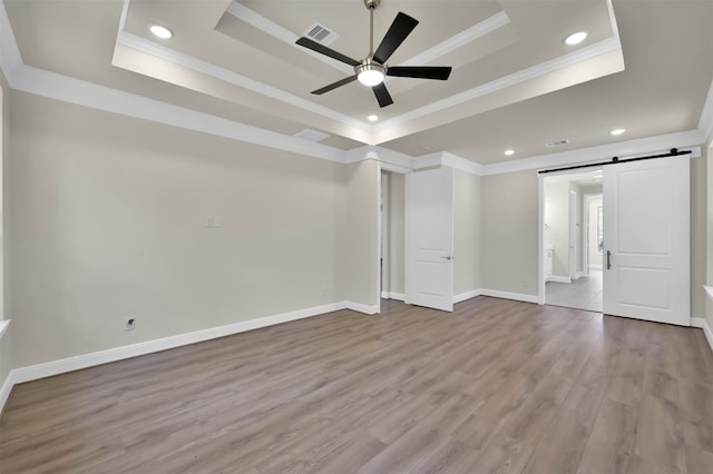 unfurnished room featuring ceiling fan, a barn door, a tray ceiling, ornamental molding, and light hardwood / wood-style floors