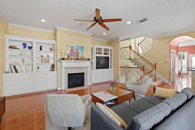 living room with built in shelves, ceiling fan, crown molding, a fireplace, and light hardwood / wood-style floors