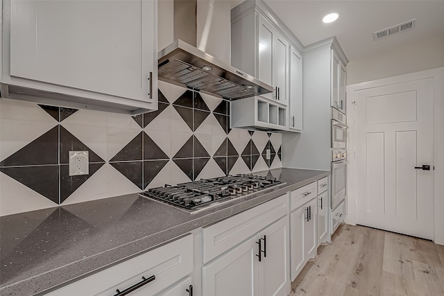 kitchen featuring wall chimney range hood, decorative backsplash, appliances with stainless steel finishes, white cabinetry, and light wood-type flooring