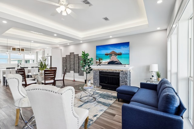 living room with ceiling fan, wood-type flooring, a premium fireplace, and a tray ceiling