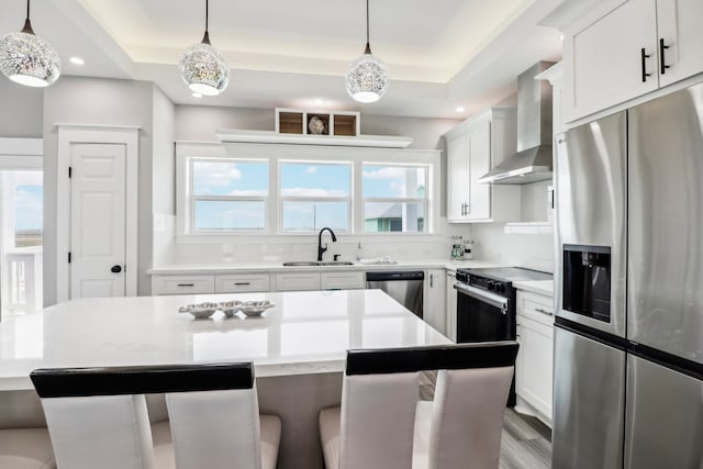 kitchen with stainless steel appliances, hanging light fixtures, a tray ceiling, and wall chimney range hood