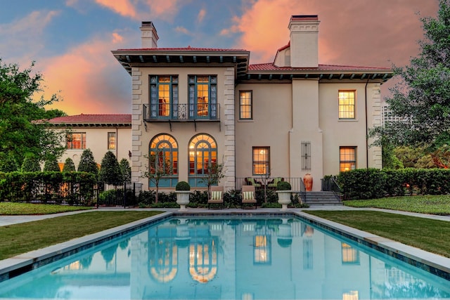 back of property at dusk featuring stucco siding, a chimney, a yard, and a fenced in pool