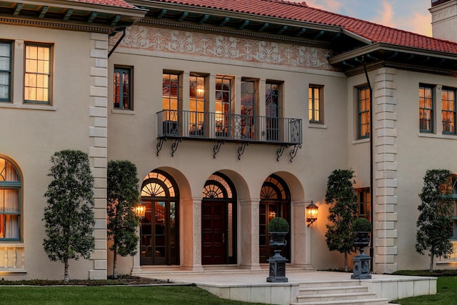back of house at dusk featuring stucco siding, a tile roof, and a balcony
