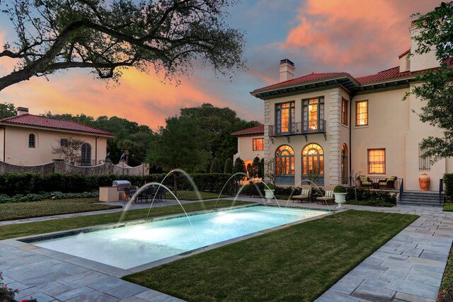 pool at dusk featuring area for grilling, a patio area, pool water feature, and a yard