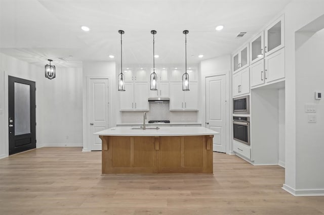 kitchen with stainless steel oven, a kitchen island with sink, built in microwave, light hardwood / wood-style floors, and white cabinetry