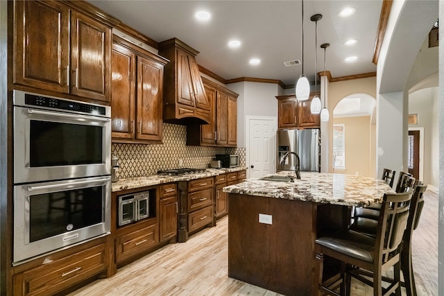 kitchen with appliances with stainless steel finishes, an island with sink, light hardwood / wood-style floors, light stone counters, and ornamental molding