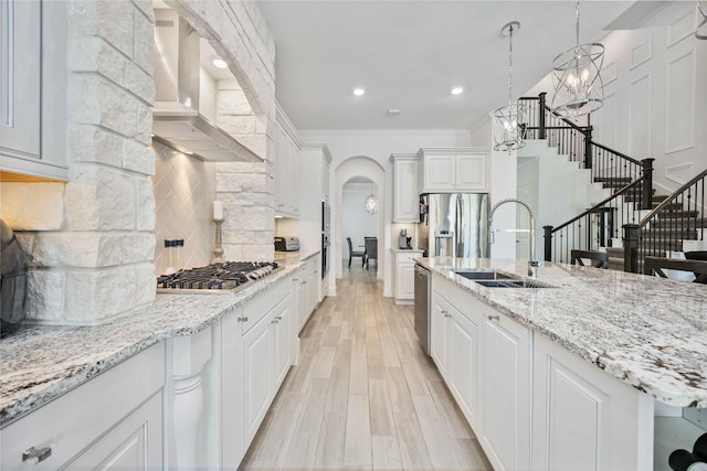 kitchen featuring white cabinets, decorative light fixtures, a kitchen island with sink, wall chimney range hood, and a sink