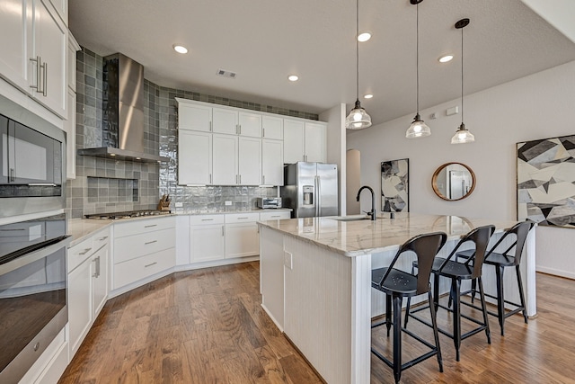 kitchen with a center island with sink, appliances with stainless steel finishes, wall chimney range hood, and white cabinets