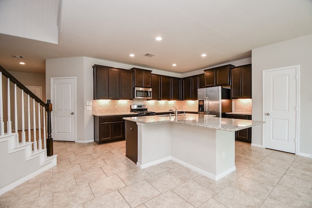 kitchen featuring a kitchen island with sink, light stone counters, stainless steel appliances, and backsplash