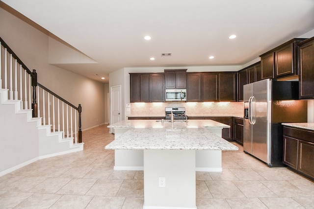 kitchen with appliances with stainless steel finishes, backsplash, dark brown cabinets, light stone counters, and a kitchen island with sink