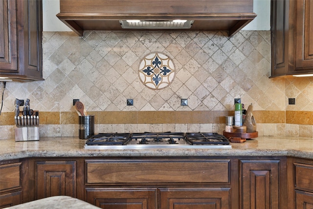 kitchen featuring dark brown cabinetry, tasteful backsplash, light stone counters, stainless steel gas stovetop, and custom range hood