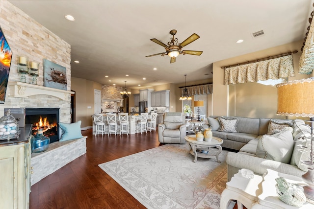 living room with dark wood-type flooring, a stone fireplace, and ceiling fan with notable chandelier