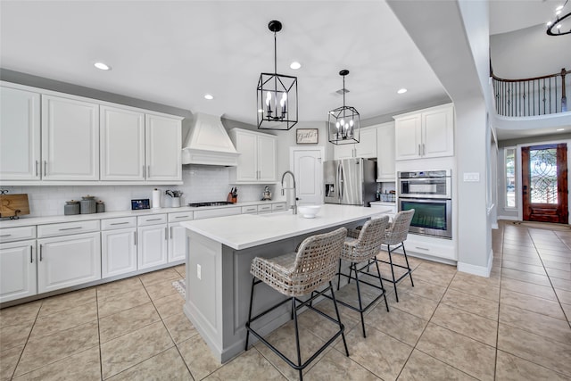 kitchen featuring custom exhaust hood, stainless steel appliances, white cabinets, a breakfast bar area, and an island with sink