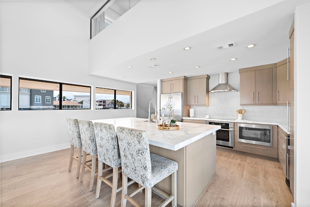 kitchen featuring appliances with stainless steel finishes, wall chimney exhaust hood, a center island with sink, light hardwood / wood-style floors, and a breakfast bar area