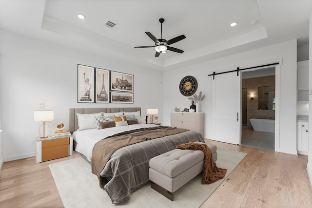 bedroom with a tray ceiling, ensuite bath, a barn door, light hardwood / wood-style floors, and ceiling fan