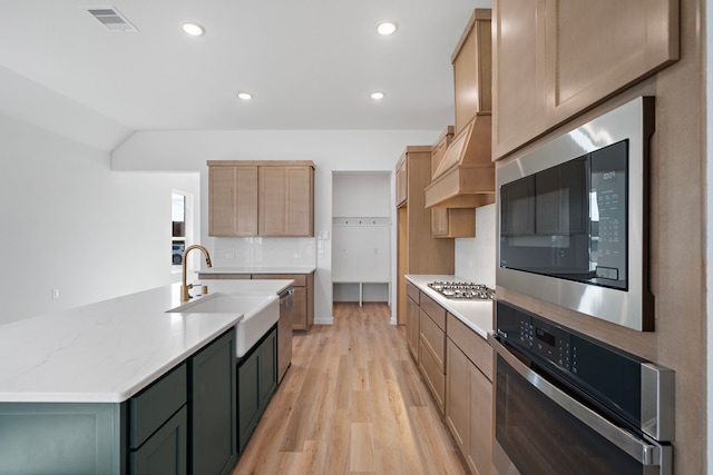 kitchen with appliances with stainless steel finishes, sink, custom exhaust hood, a kitchen island with sink, and light wood-type flooring