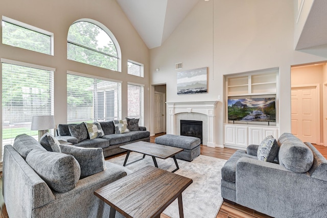 living room featuring a tiled fireplace, high vaulted ceiling, light hardwood / wood-style flooring, and built in shelves