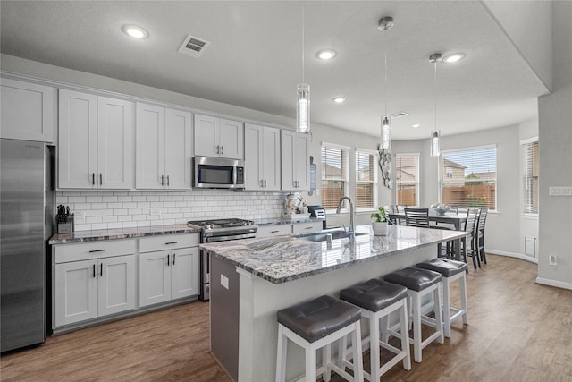 kitchen featuring an island with sink, hanging light fixtures, stainless steel appliances, wood-type flooring, and white cabinets