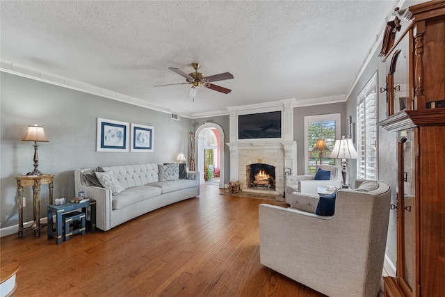 living area featuring arched walkways, ceiling fan, a stone fireplace, a textured ceiling, and wood finished floors