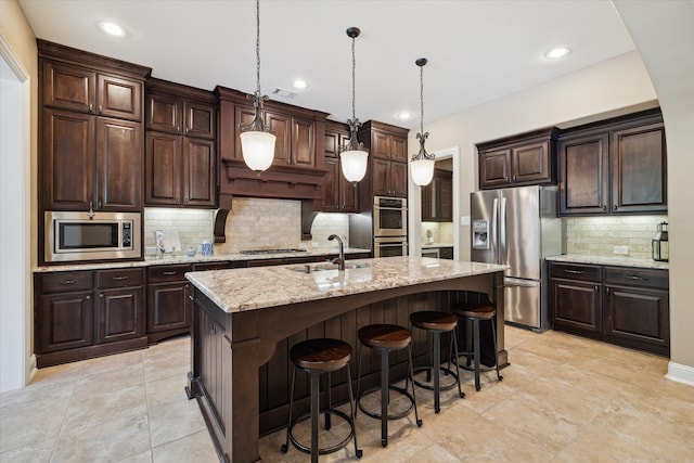 kitchen with appliances with stainless steel finishes, dark brown cabinets, decorative light fixtures, and a kitchen island with sink