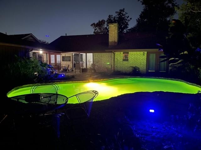 back of house at night featuring an outdoor pool, brick siding, a patio, and a chimney