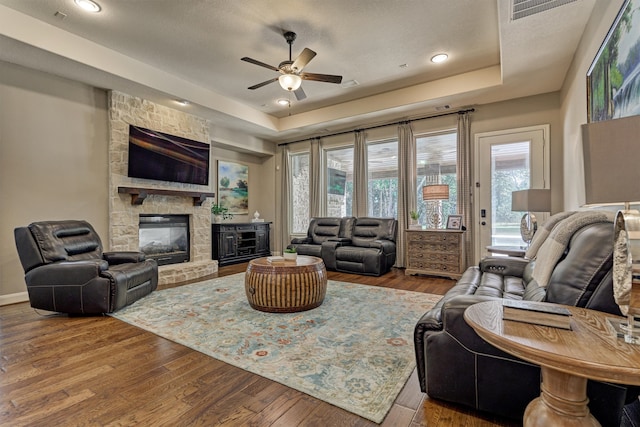 living room featuring ceiling fan, a raised ceiling, a textured ceiling, wood-type flooring, and a stone fireplace