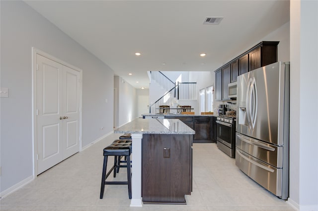 kitchen with light stone countertops, dark brown cabinetry, a center island, a kitchen bar, and appliances with stainless steel finishes