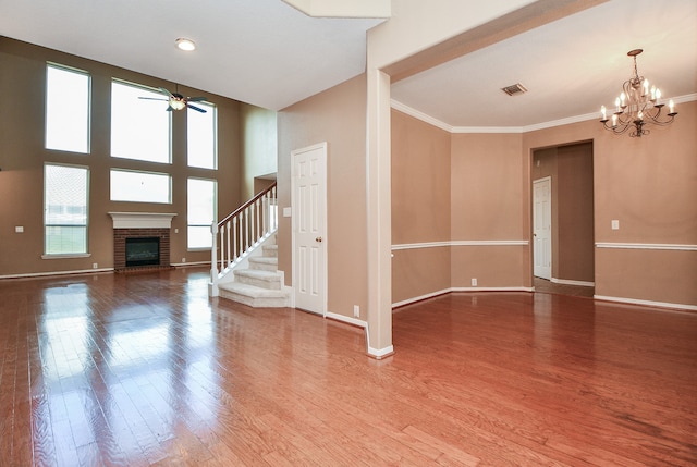 unfurnished living room featuring a high ceiling, hardwood / wood-style flooring, ornamental molding, a brick fireplace, and ceiling fan with notable chandelier